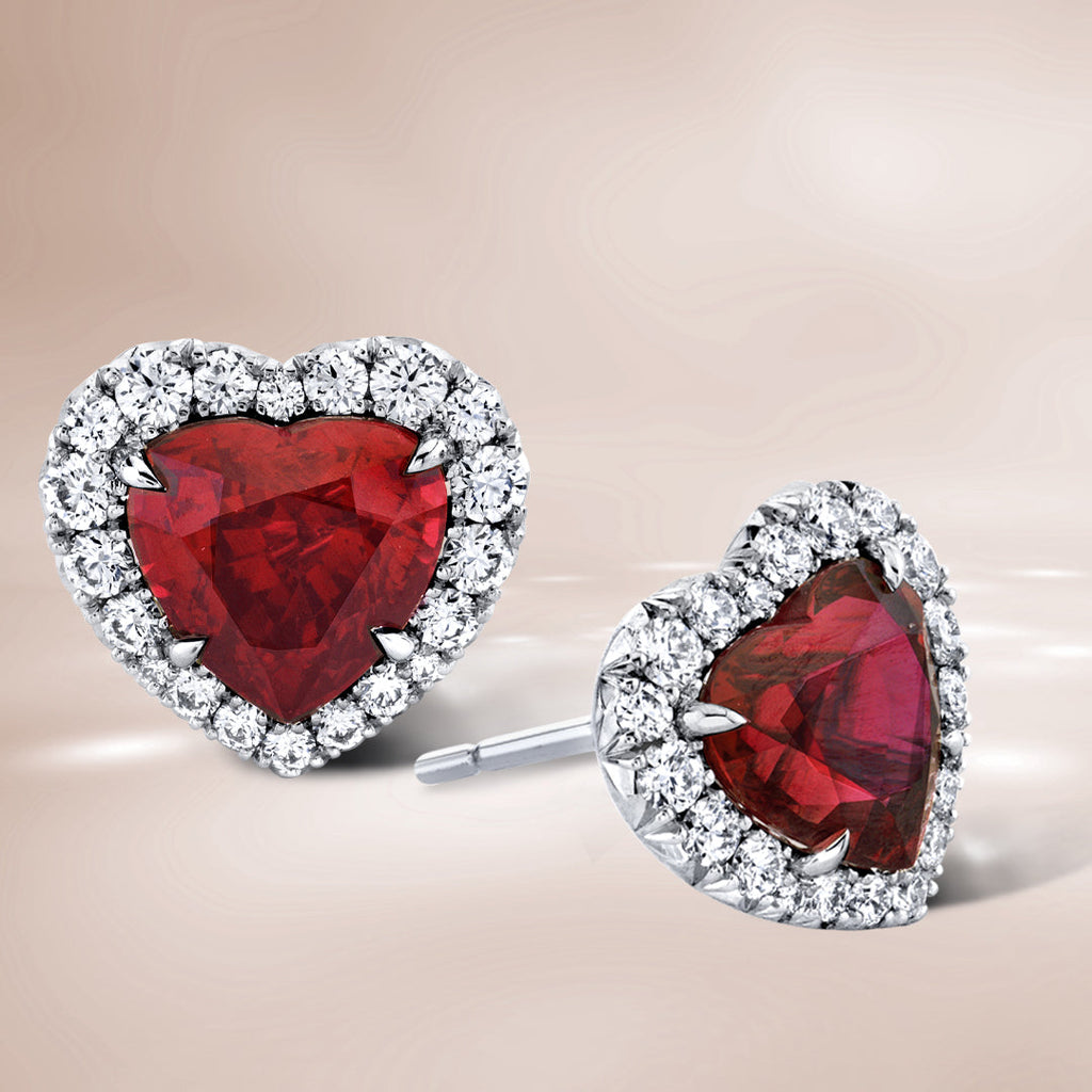 Heart shaped ruby stud earrings with diamond halos on a brown background.