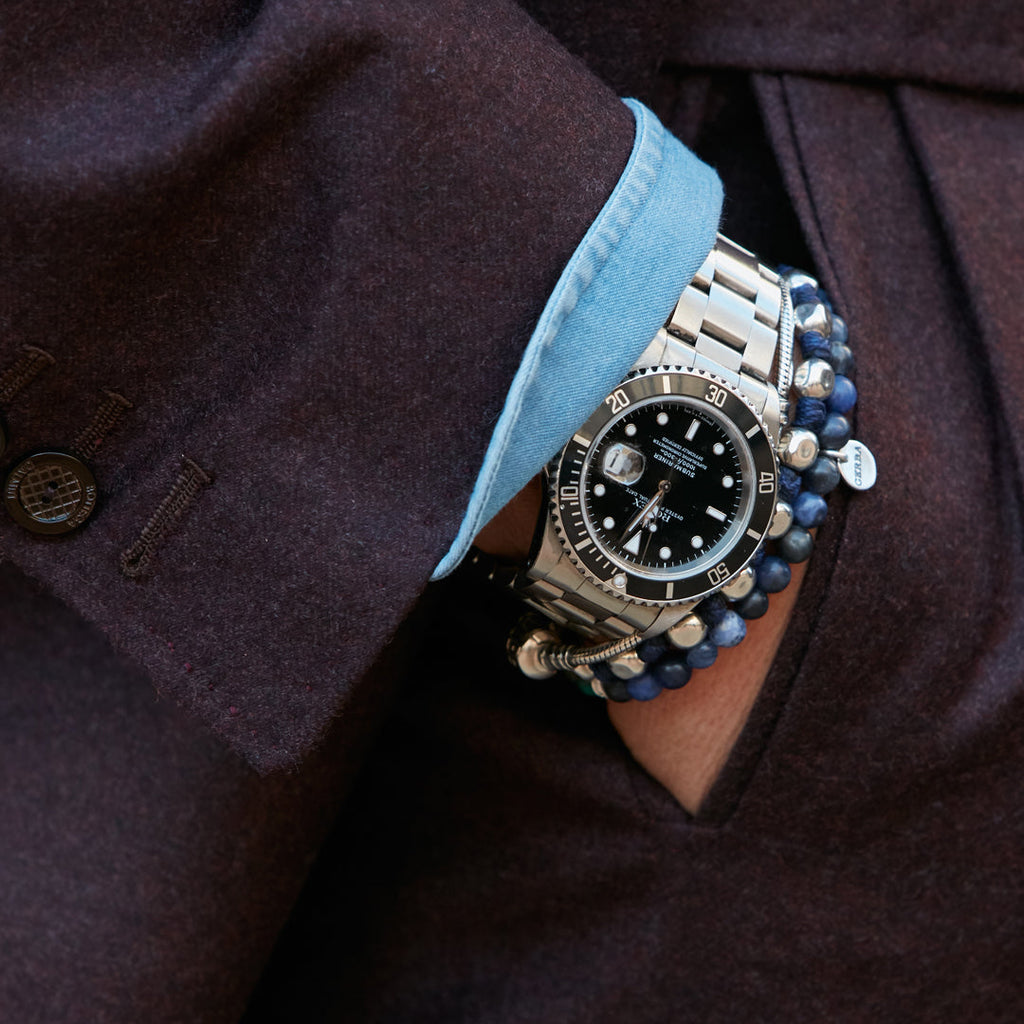 Man with hand in suit pocket wearing a watch and beaded bracelets.